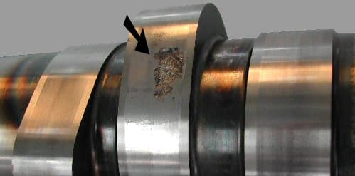 A camshaft surface showing pitting and spalling, with the affected area marked by an arrow